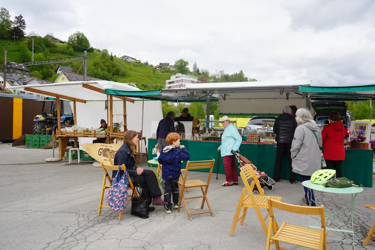 Man sieht Leute an einem Markt. Es gibt auch Tische zum Verweilen. Dort sitzen Leute.