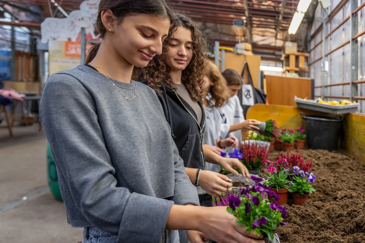 Am Pflanztisch durften die Schülerinnen nach einer kurzen Anleitung selber Hand anlegen und einen kleinen Korb mit Blumen bepflanzen. Man sieht junge Frauen, wie sie Blumen pflanzen.