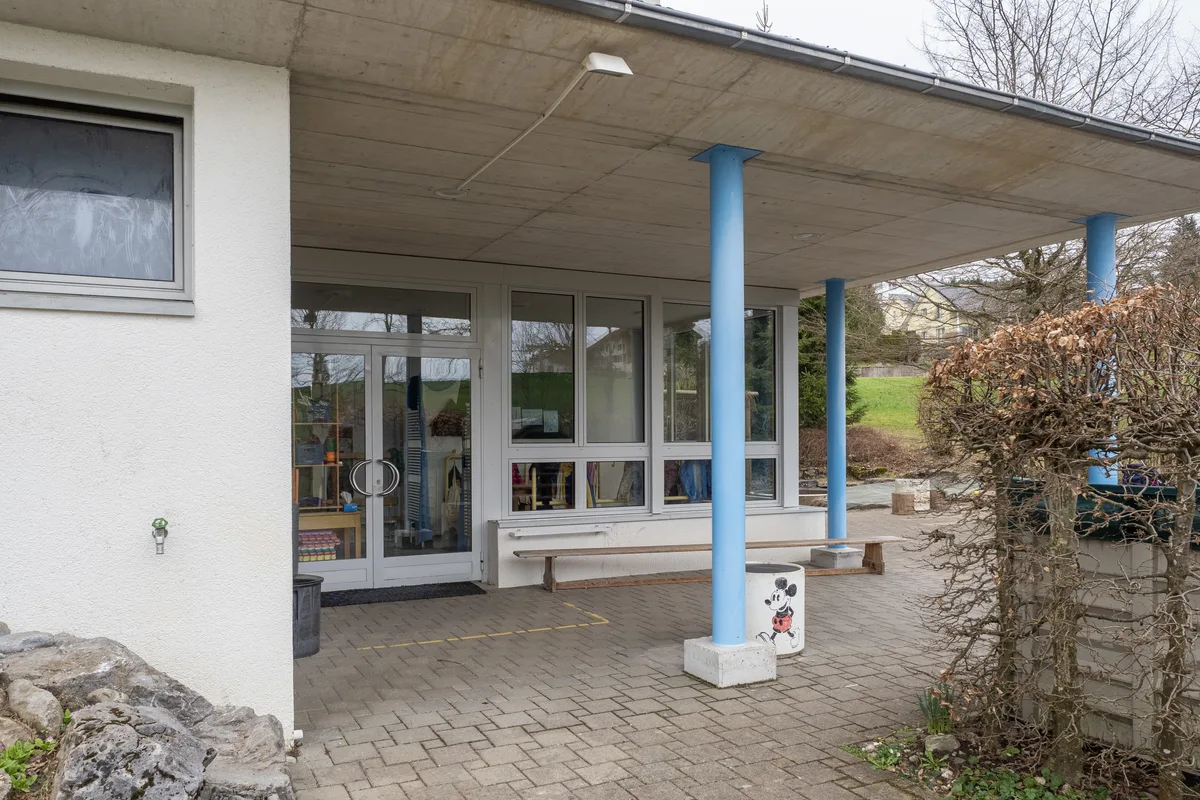 Hinter dem bisherigen Eingang wird künftig das Materiallager sein. Man sieht den gedeckten Eingang zu einem Gebäude, mit Glastür und blauen Säulen.