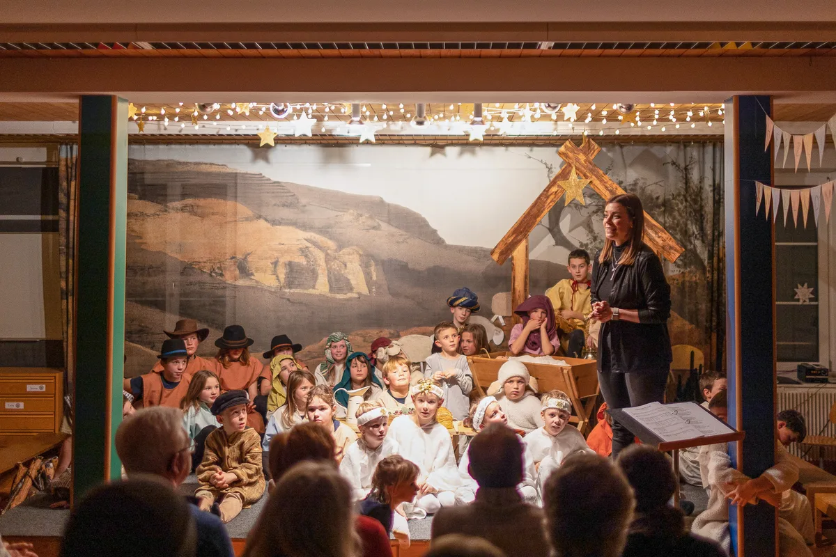 Sarah Köhler stellt sich als neue Schulleiterin vor. Man sieht eine Frau, die auf der Bühne steht. Hinter hier sind die Kinder vom Weihnachtsspiel versammelt.