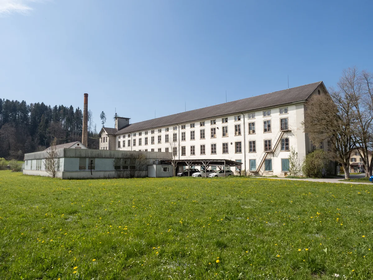 Auf der Wiese hinter der Fabrik sind zahlreiche Neubauten geplant. Der hellblaue Anbau wird abgerissen. (Archiv) Man sieht eine Wiese, Fabrik und Kamin.