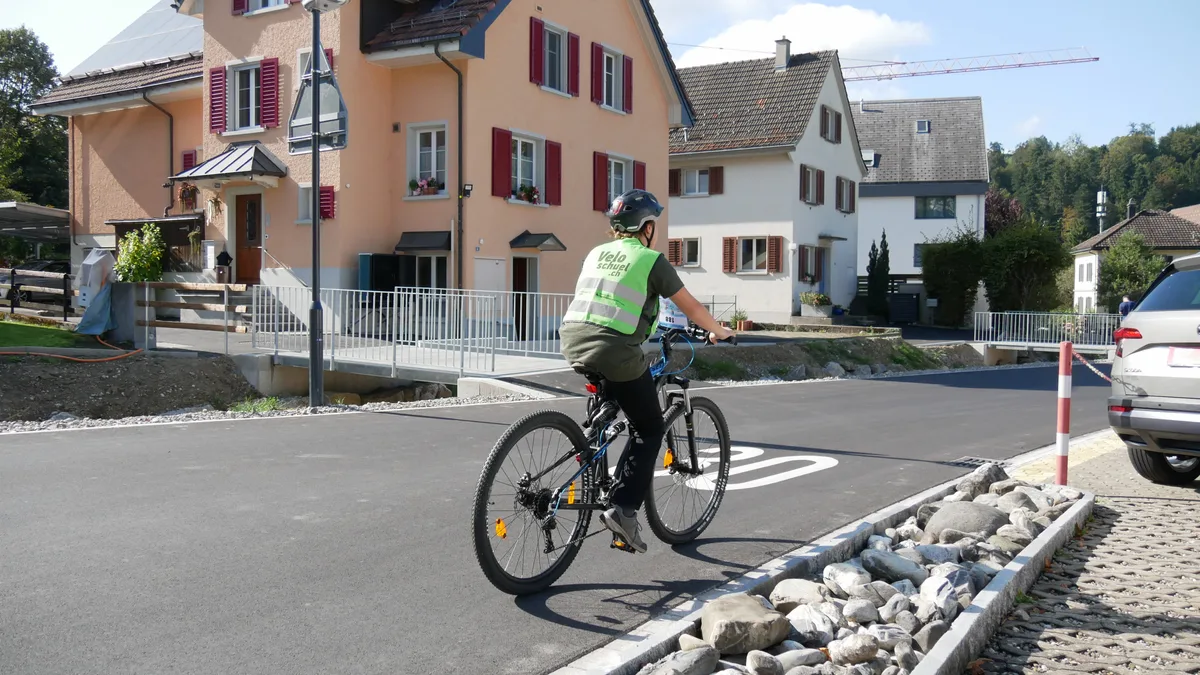 Man sieht einen Jungen mit Warnweste, der mit dem Velo über eine Strasse fährt.