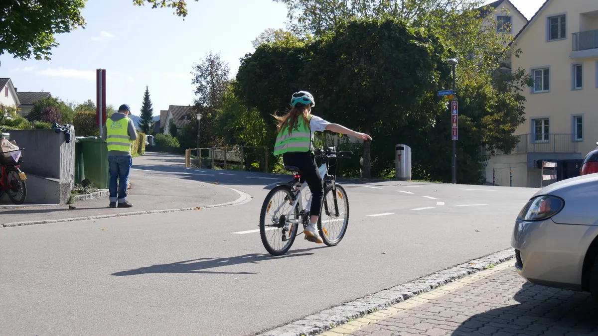 Man sieht ein Mädchen mit Warnweste, das mit dem Velo über eine Strasse fährt.