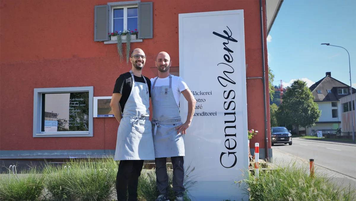 Foto von zwei Männern mit grauer Schürze und Glatze vor einem Verkaufslokal.