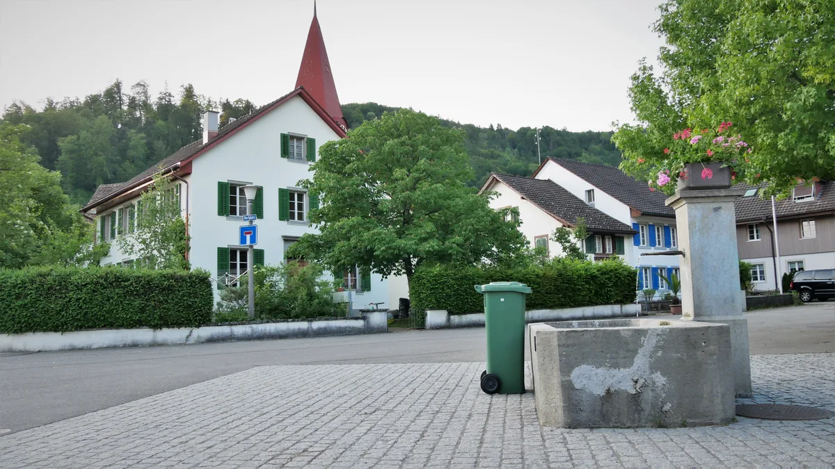 Man sieht einen Dorfplatz mit einer Kirche im Hintergrund und einem Dorfbrunnen am rechten Bildrand.