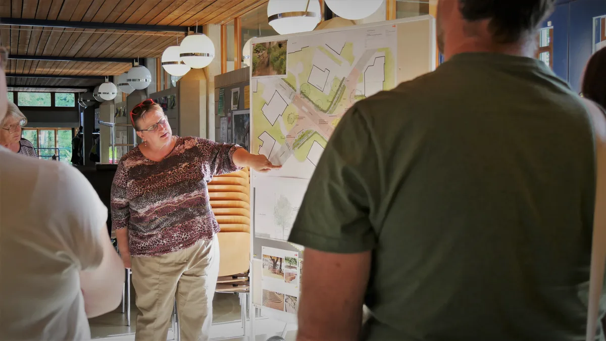 Eine Frau mittleren Alters zeigt mit dem Arm auf einen schematischen Plan, während andere ihr zuhören.