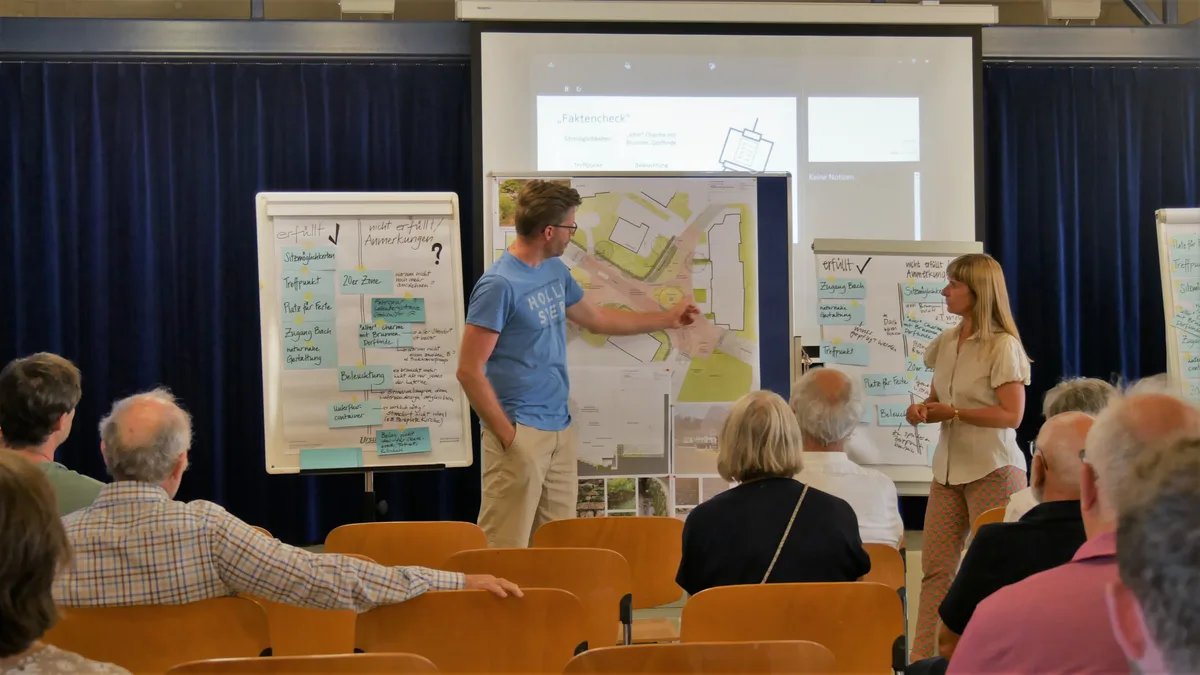 Ein Mann in einem blauen Shirt zeigt auf einen schematischen Plan. Eine Frau mit gemusterten Hosen und weisser Bluse diskutiert mit.