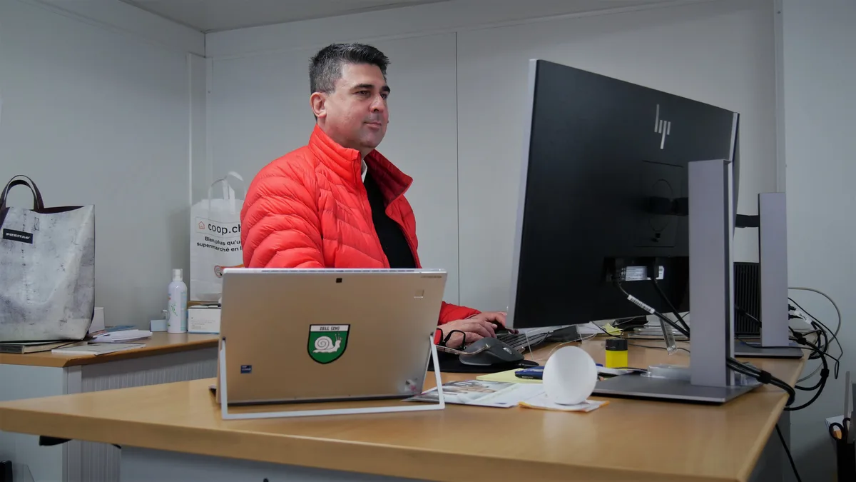 Man sieht den Gemeindeschreiber von Zell, Erkan Metschli-Roth, mit einer roten Winterjacke, an seinem Arbeitsplatz im provisorischen Gemeindehaus Zell.