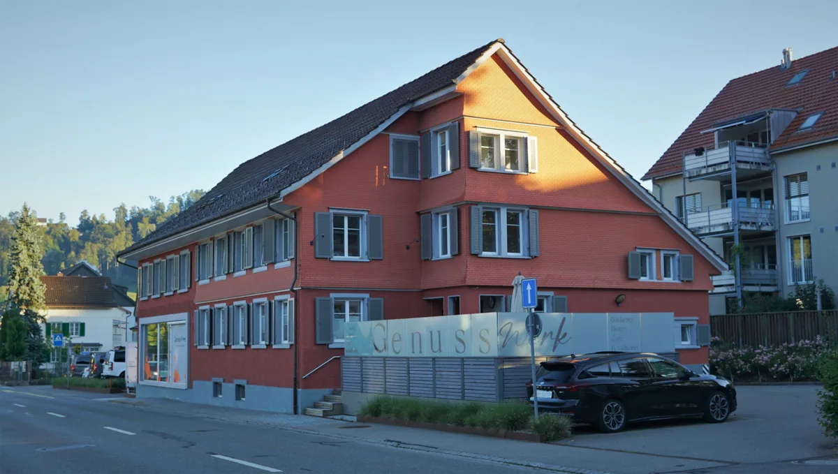 Man sieht ein rosarotes Haus mit einem Verkaufslokal und einer Terrasse an einer Hauptstrasse.