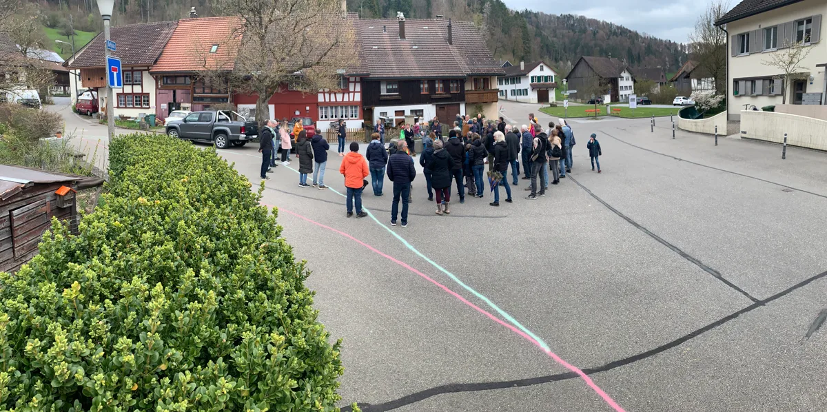 Man sieht eine Gruppe Menschen, die sich auf einem Dorfplatz versammelt hat.