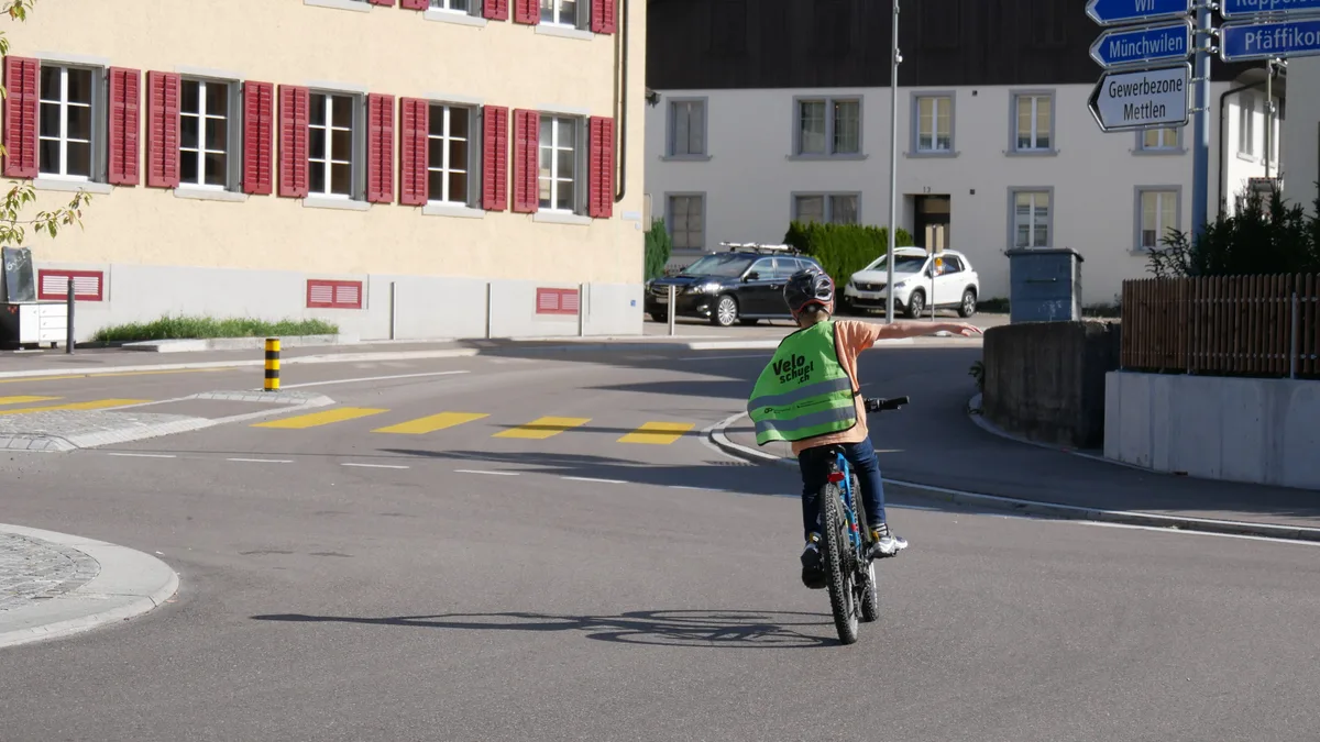 Man sieht einen Jungen mit Warnweste, der mit dem Velo über eine Strasse fährt.