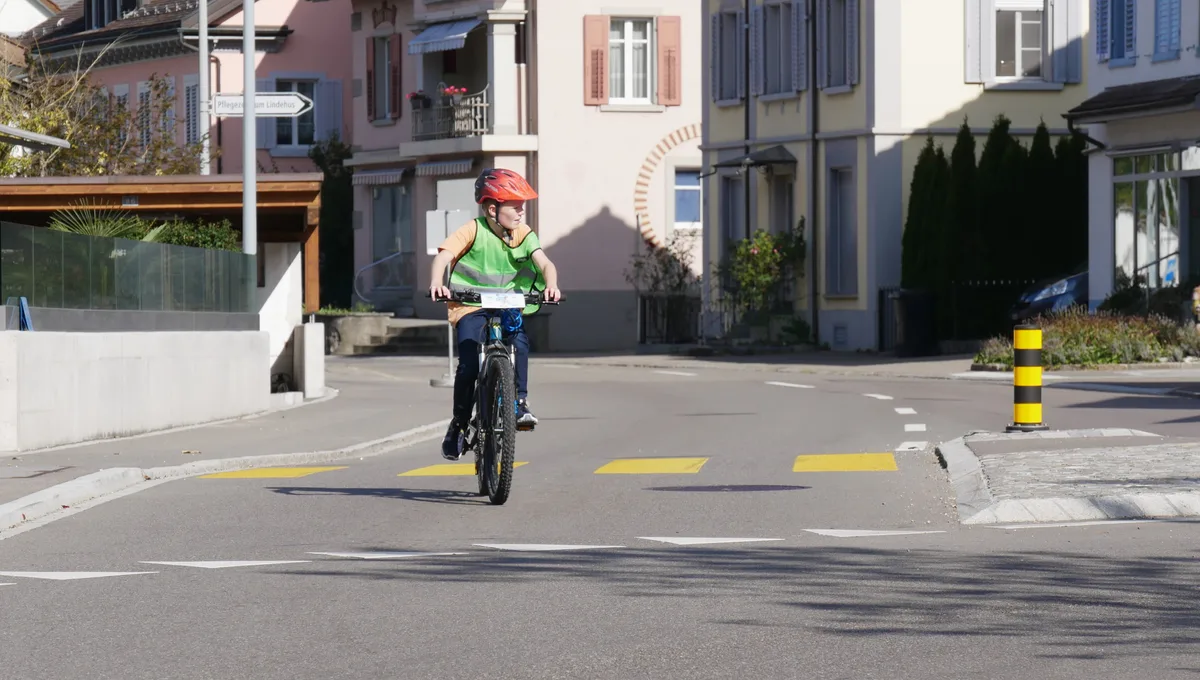 Man sieht einen Jungen mit Warnweste, der mit dem Velo über eine Strasse fährt.