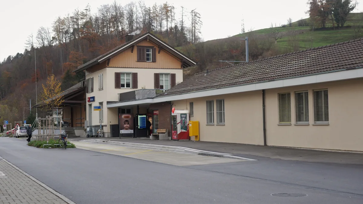 Man sieht das Stationsgebäude des Bahnhofs Kollbrunn sowie die Bushaltestelle davor.