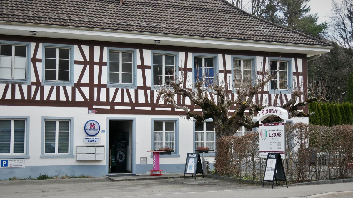 Man sieht ein altes Fachwerkhaus mit roten Balken und einem Garten an einer vielbefahrenen Strasse. Es handelt sich um ein Restaurant.