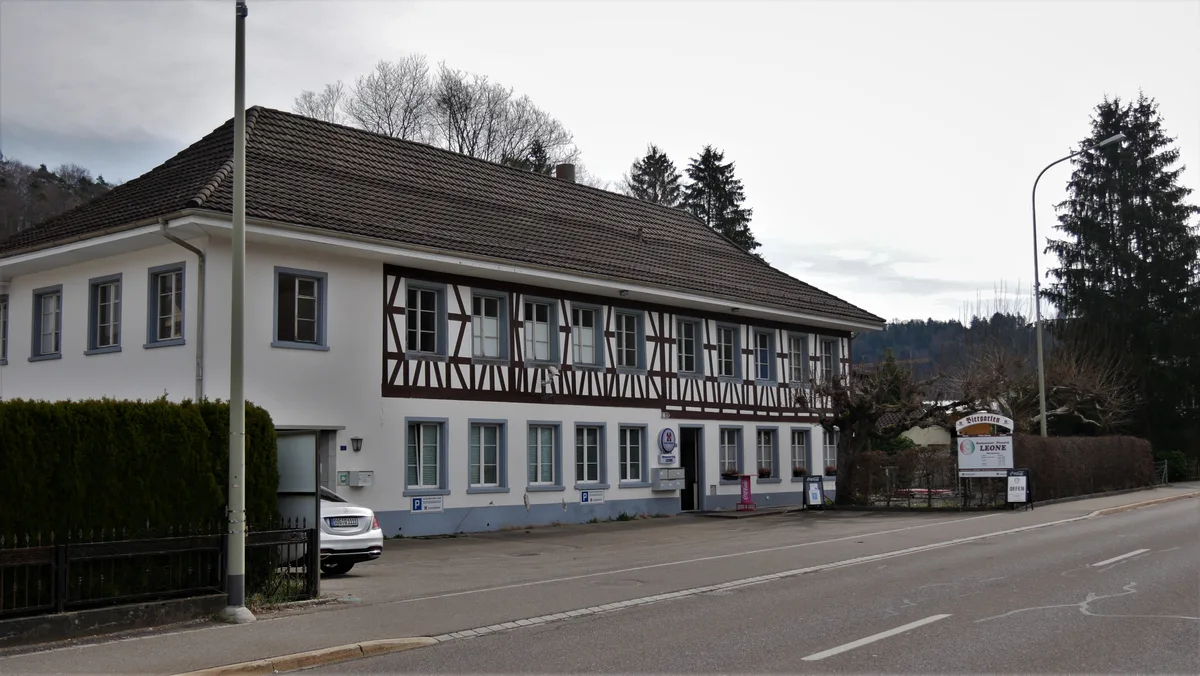 Man sieht ein altes Fachwerkhaus mit roten Balken und einem Garten an einer viel befahrenen Strasse. Es handelt sich um ein Restaurant.