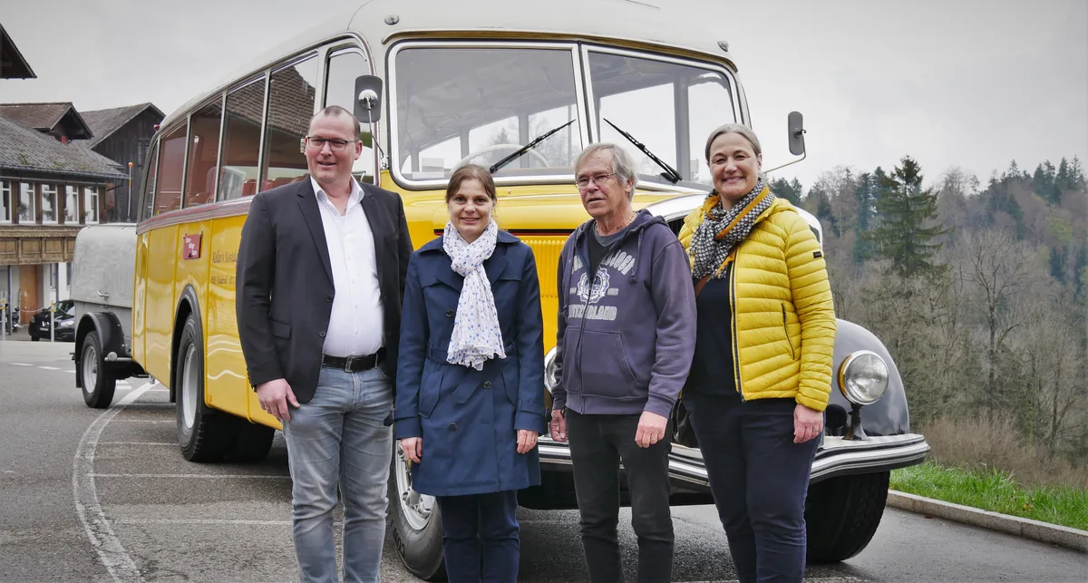Man sieht vier Menschen, die vor einem alten Postauto für ein Foto posieren.