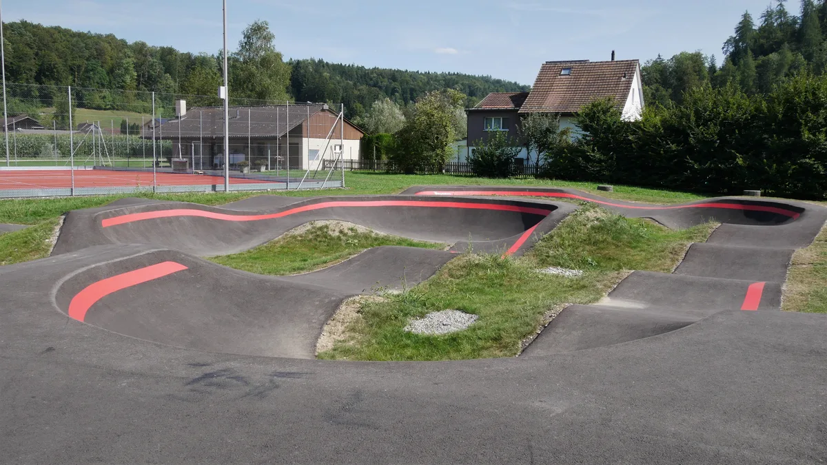 Es ist eine asphaltierte Pumptrack-Strecke mit farbigen Markierungen zu sehen.