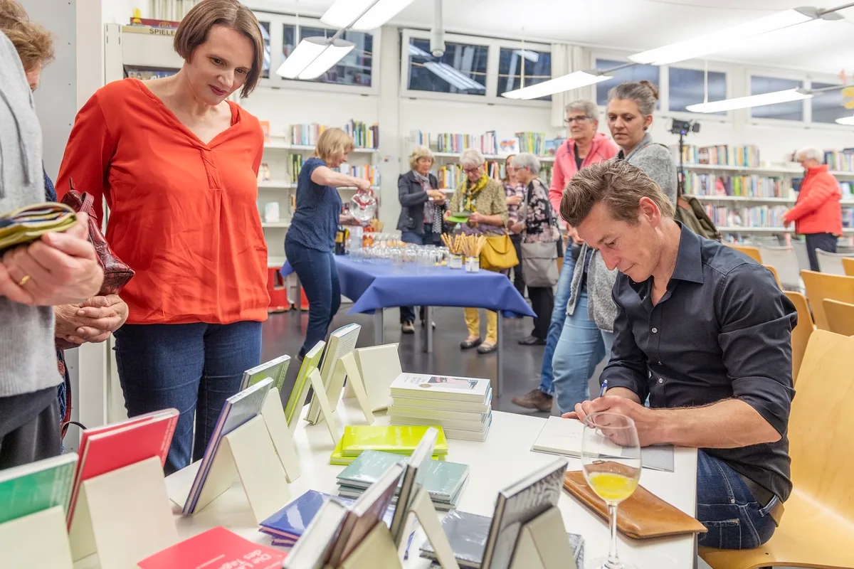 Man sieht Personen, die an einem Büchertisch warten. Ein Mann signiert ein Buch.