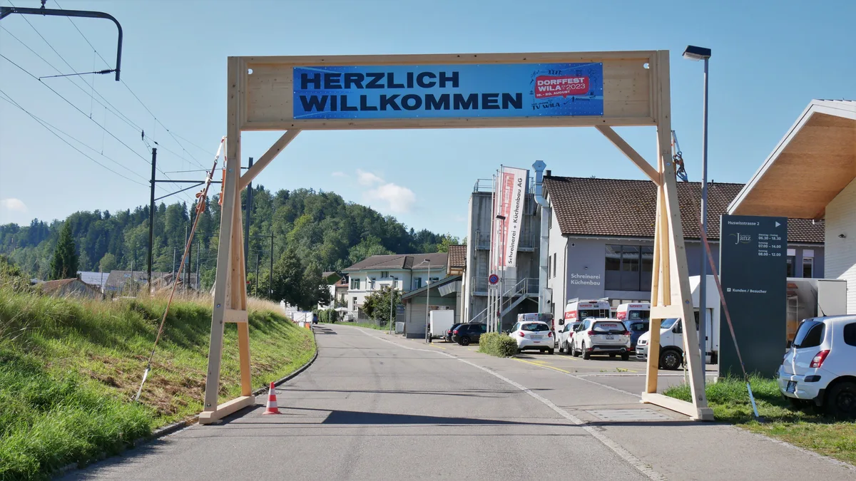 Ein hölzerner Torbogen steht im Dorfzentrum. Darauf ist ein Plakat montiert.