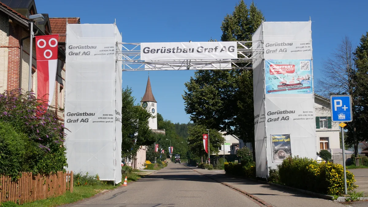 Ein weisser Torbogen steht im Dorfzentrum. Darauf sind Plakate montiert.