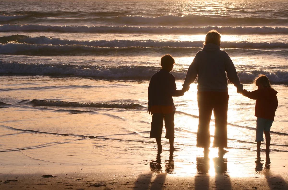 Man sieht eine Frau, die zwei Kinder an der Hand hält und mit ihnen am Strand steht.