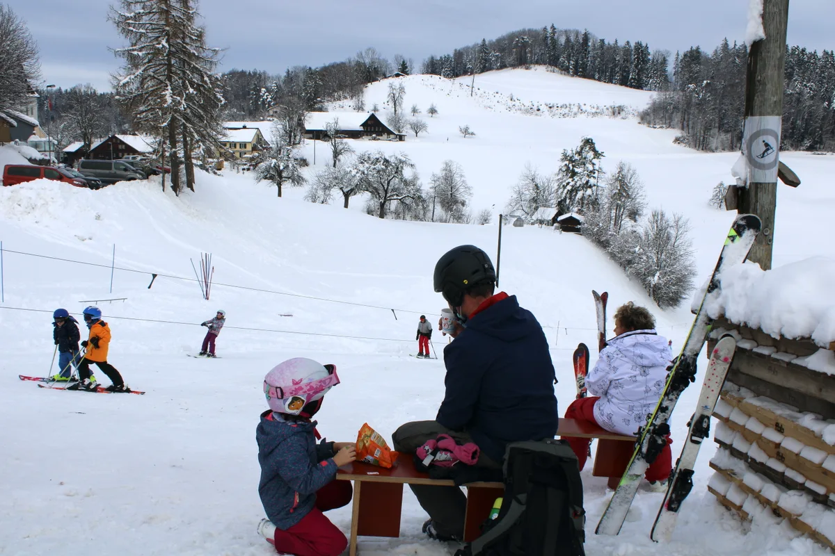 Man sieht Leute an einem Skilift und Vater und Tochter auf einer Bank.