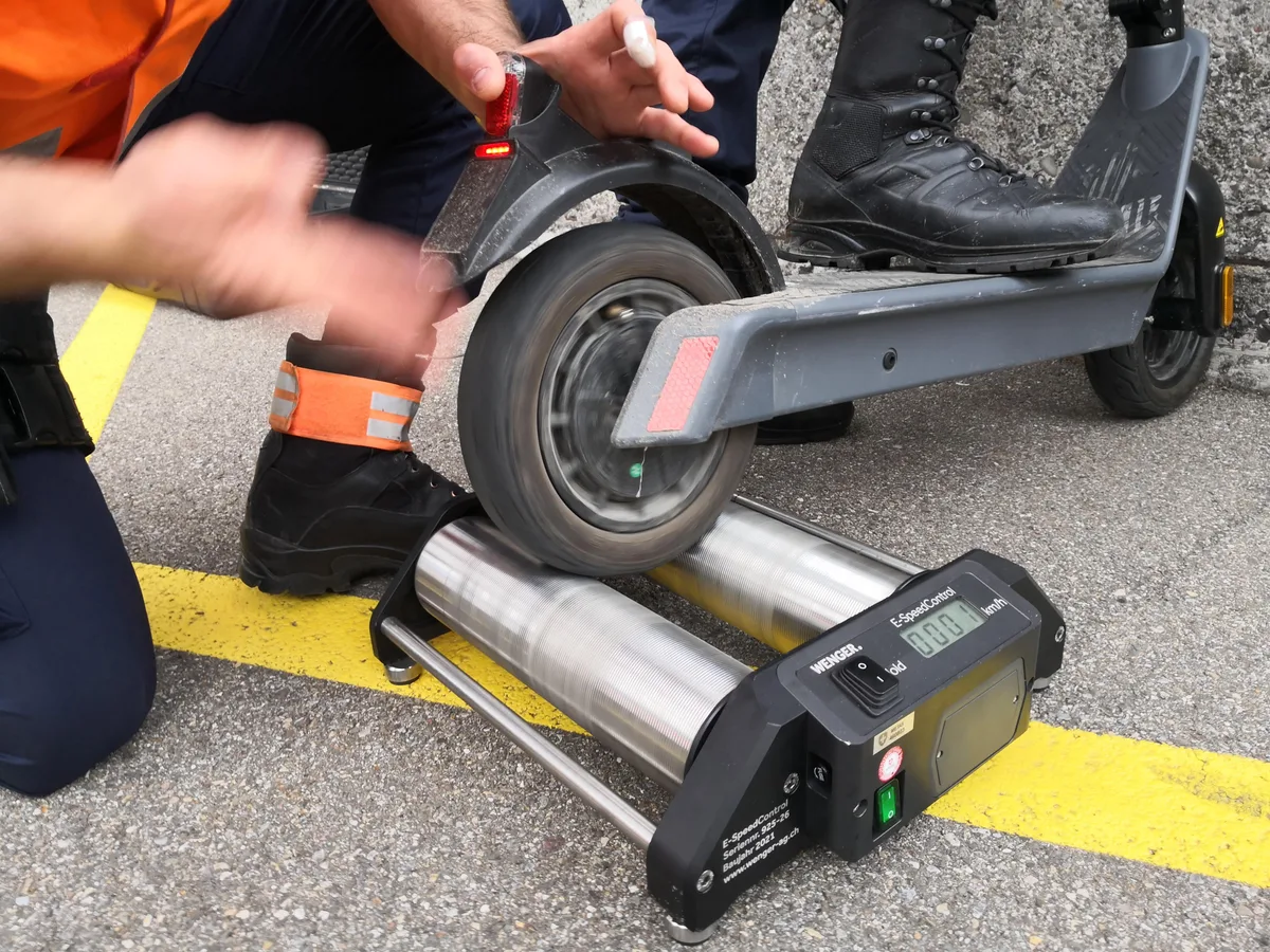 Man sieht einen Teil eines knienden Menschen. Dieser hält gerade das hintere Rad eines E-Scooters auf ein Gerät mit Rollen, mit dem er die Geschwindigkeit messen kann.