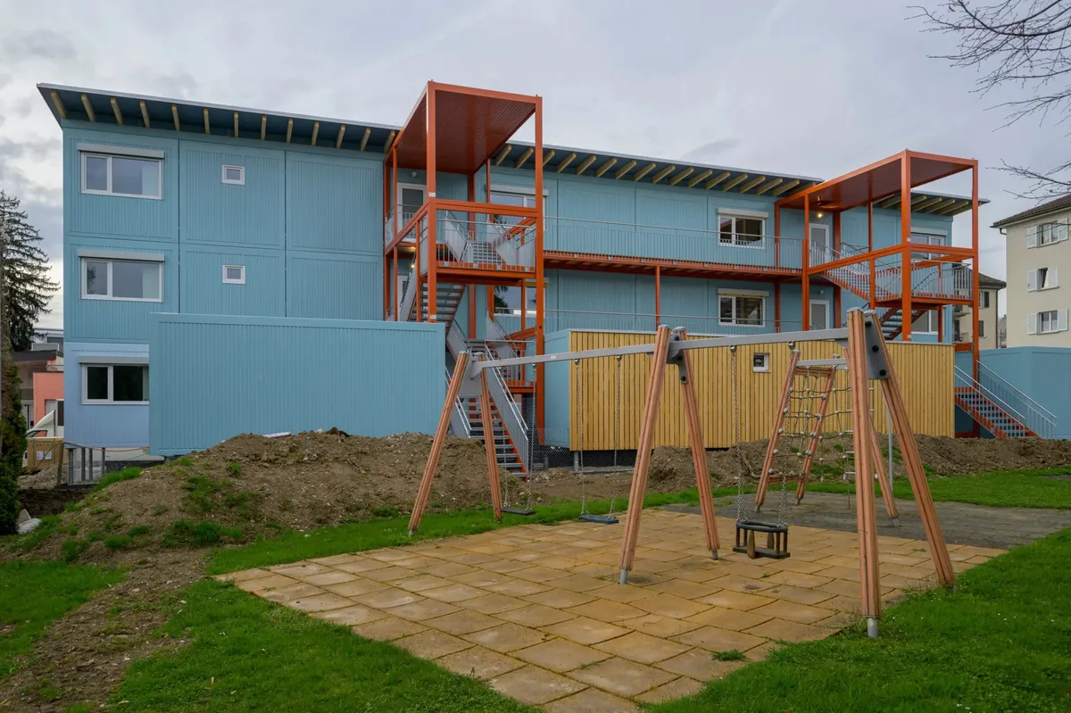 Man sieht einen Spielplatz. Im Hintergrund befindet sich ein Containerbau.