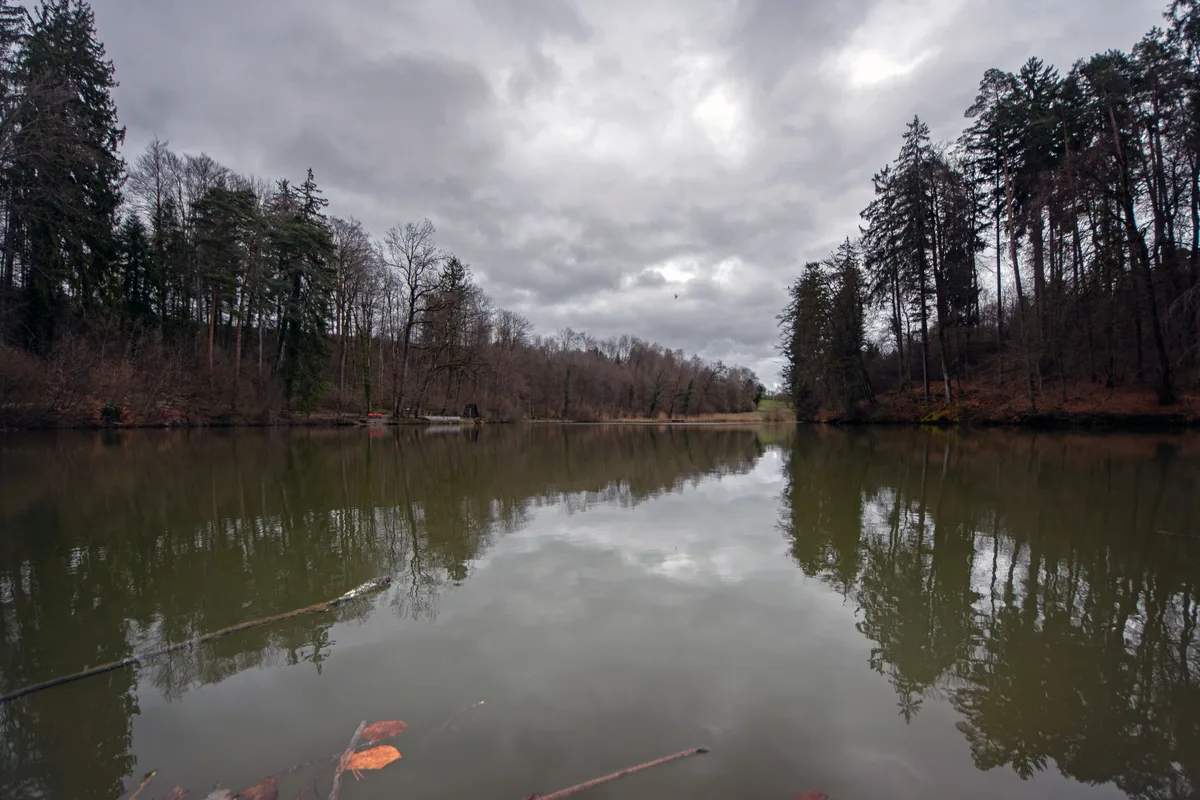 Am späten Nachmittag desselben Tages, etwa um 17 Uhr: Nun ist es beinahe windstill. Zuvor war ein kurzer, aber intensiver Graupelschauer über den Weiher gezogen. Ein Teich mit grauem Himmel.