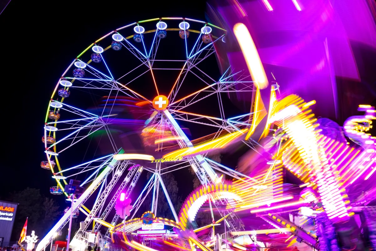 Bei Nacht grell beleuchtete Fahrgeschäfte. Im Hintergrund ist ein Riesenrad zu sehen.