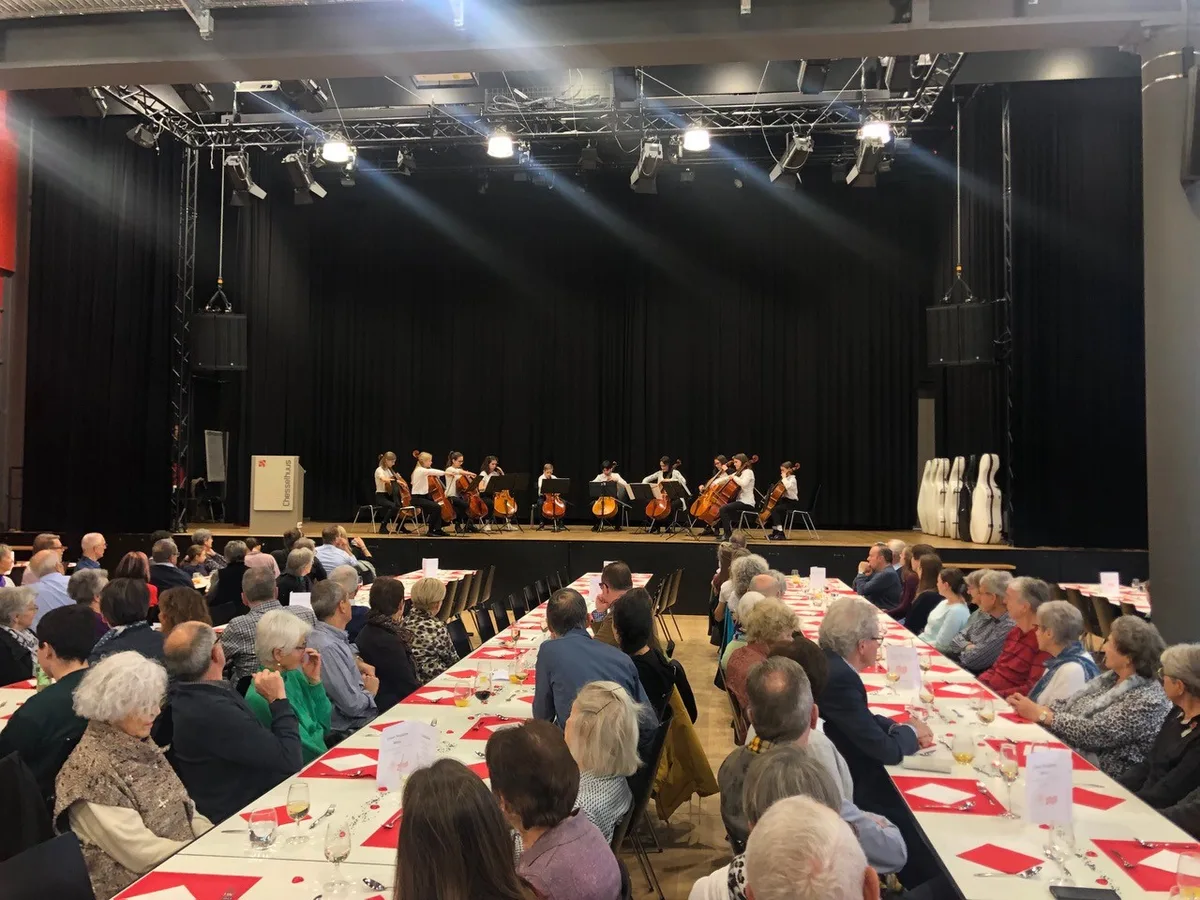 Die Cello-Kracher auf der Bühne des Pfiffiger Chesselhuus. Man sieht Leute in einem Saal mit Tischen, die einem Cello-Ensemble auf der Bühne zuhören.