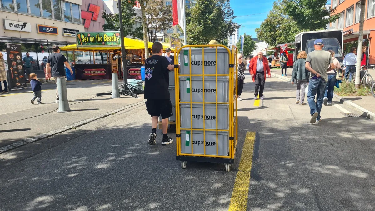Gut für die Umwelt, etwas mühsam im Transport: die Mehrwegbecher von Cup & More. Ein Mann zieht einen Wagen, bepackt mit Kisten die mit Cup n More beschriftet sind.