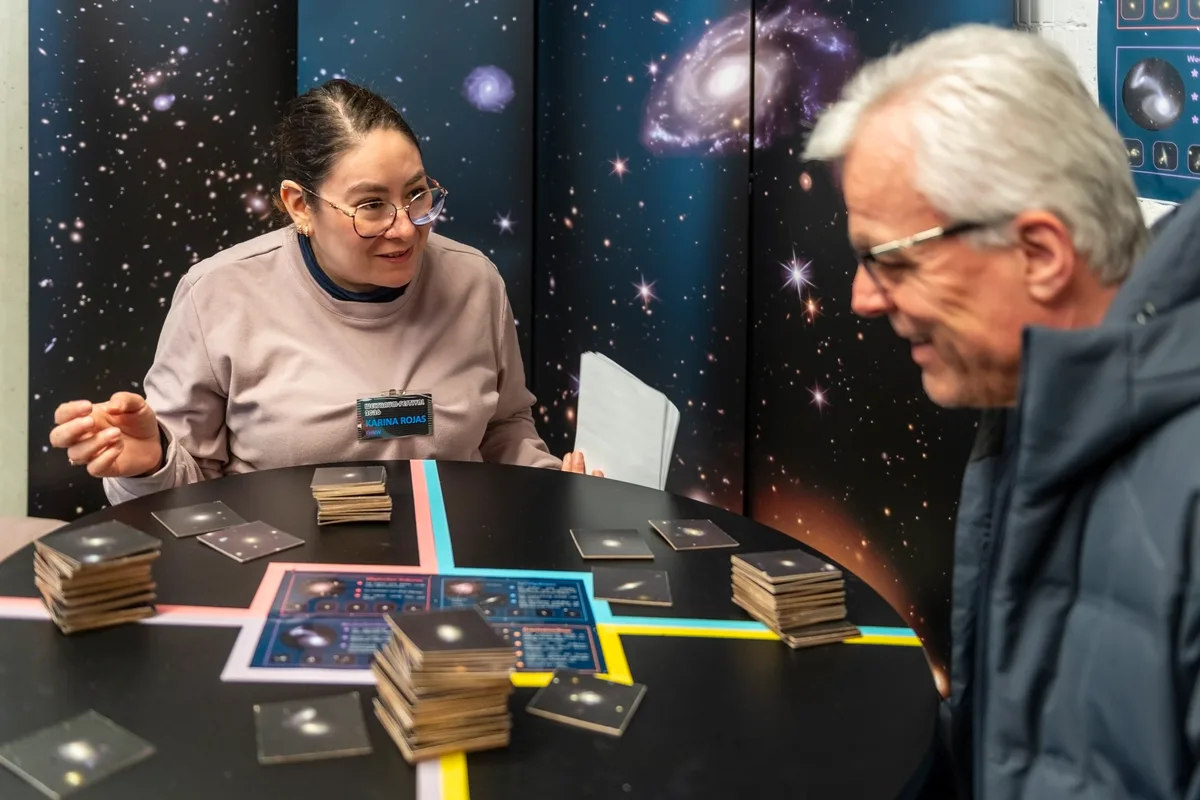 Karina Rojas und ein Mann sitzen an einem Tisch mit Kärtchen. Sie erklärt ihm etwas.