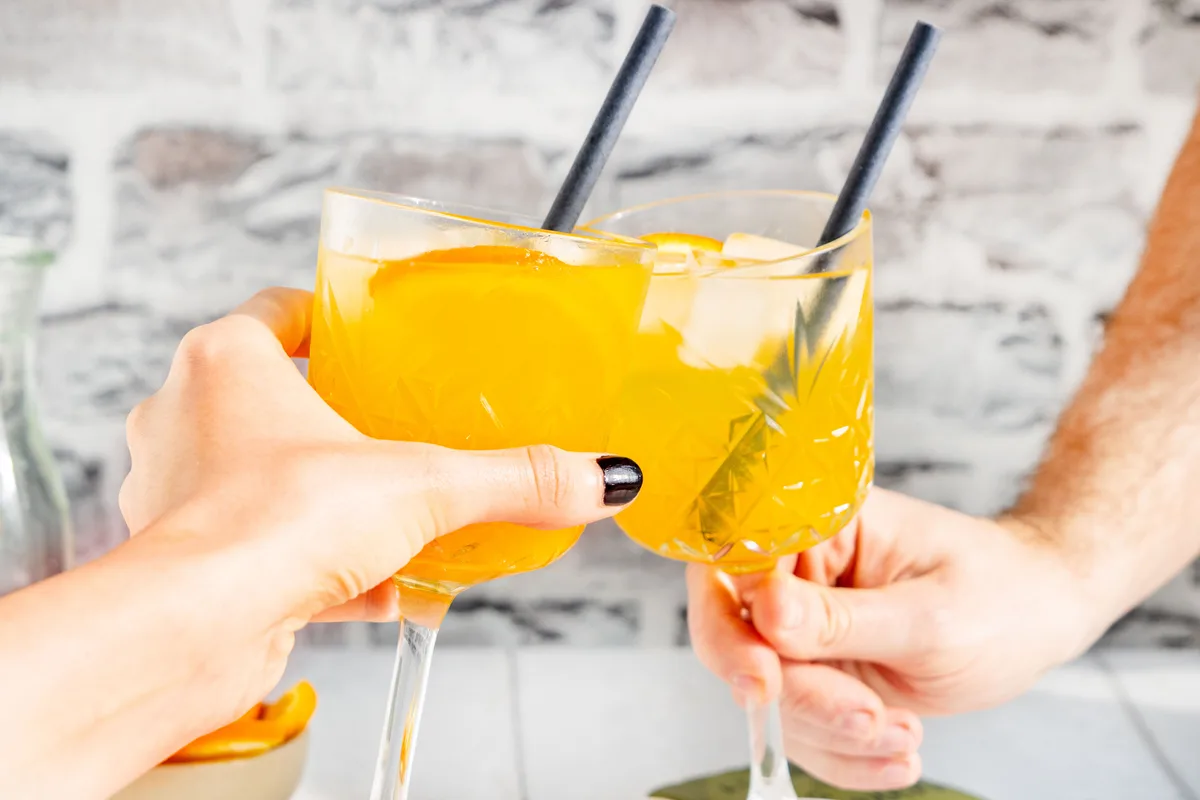 Auch mit einem Mocktail lässt es sich anstossen. Ob im Drink Alkohol drin ist, lässt sich oft überhaupt nicht sagen. (Symbolbild) Man sieht zwei Gläser mit einem orangen Getränk und Personen, die anstossen.