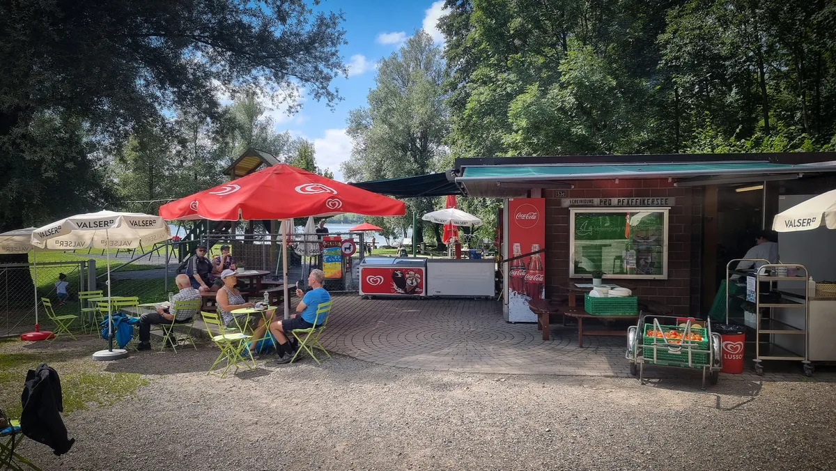 Rechts ein Kioskgebäude, links sitzen Mensch an Tischen unter Sonnenschirmen. 