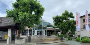 29605545-3 Oberland Märt Oberlandmärt Migros Wetzikon am 14. Juli 2021 Eingangsbereich mit vier Platanen und dem Buswartehäuschen. Drei der vier Bäume sollen für die Neugestaltung des Platzes weg.