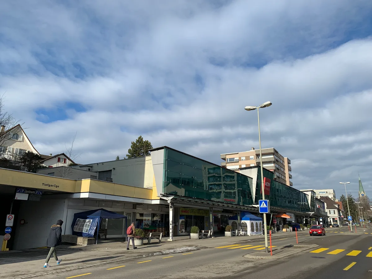 Ein Einkaufszentrum in Wetzikon und davor Fussgänger und ein Auto.  