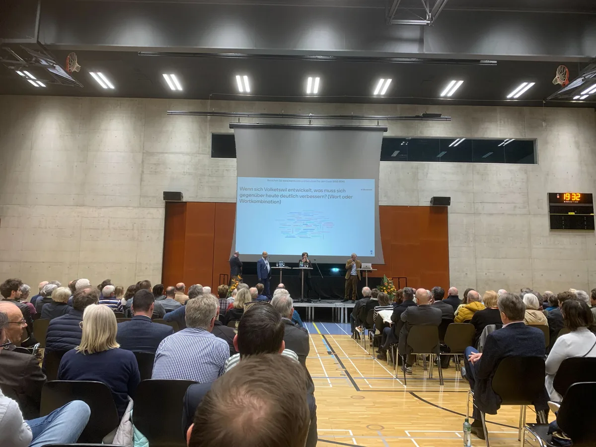 Viele Leute sitzen in einer Turnhalle auf Stühlen und warten auf den Beginn einer Info-Veranstaltung.