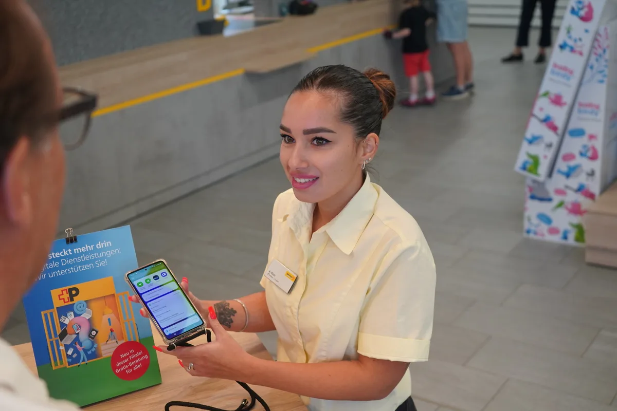 Eine junge Postangestellte hält ein Smartphone in der Hand und erklärt einem Kunden etwas.