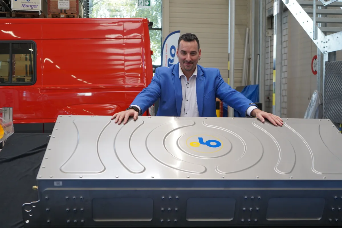 Ein Mann in blauem Jackett und weissem Hemd steht hinter einer grossen silbernen Kiste, die eine Autobatterie enthält.