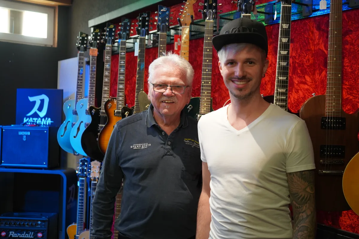 Ein älterer und ein junger Mann stehen in einem Musikgeschäft vor einer Wand, an der zahlreiche Gitarren hängen.