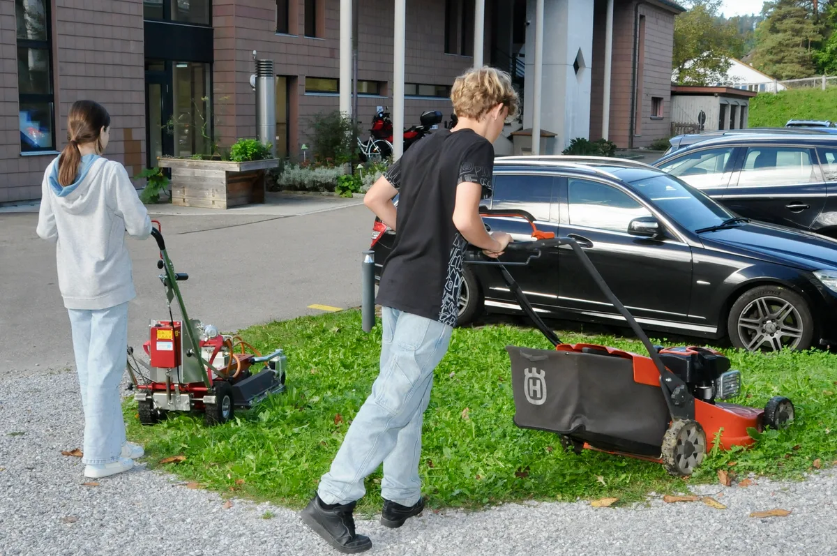Man sieht zwei Jugendliche mit dem Rasenmäher.