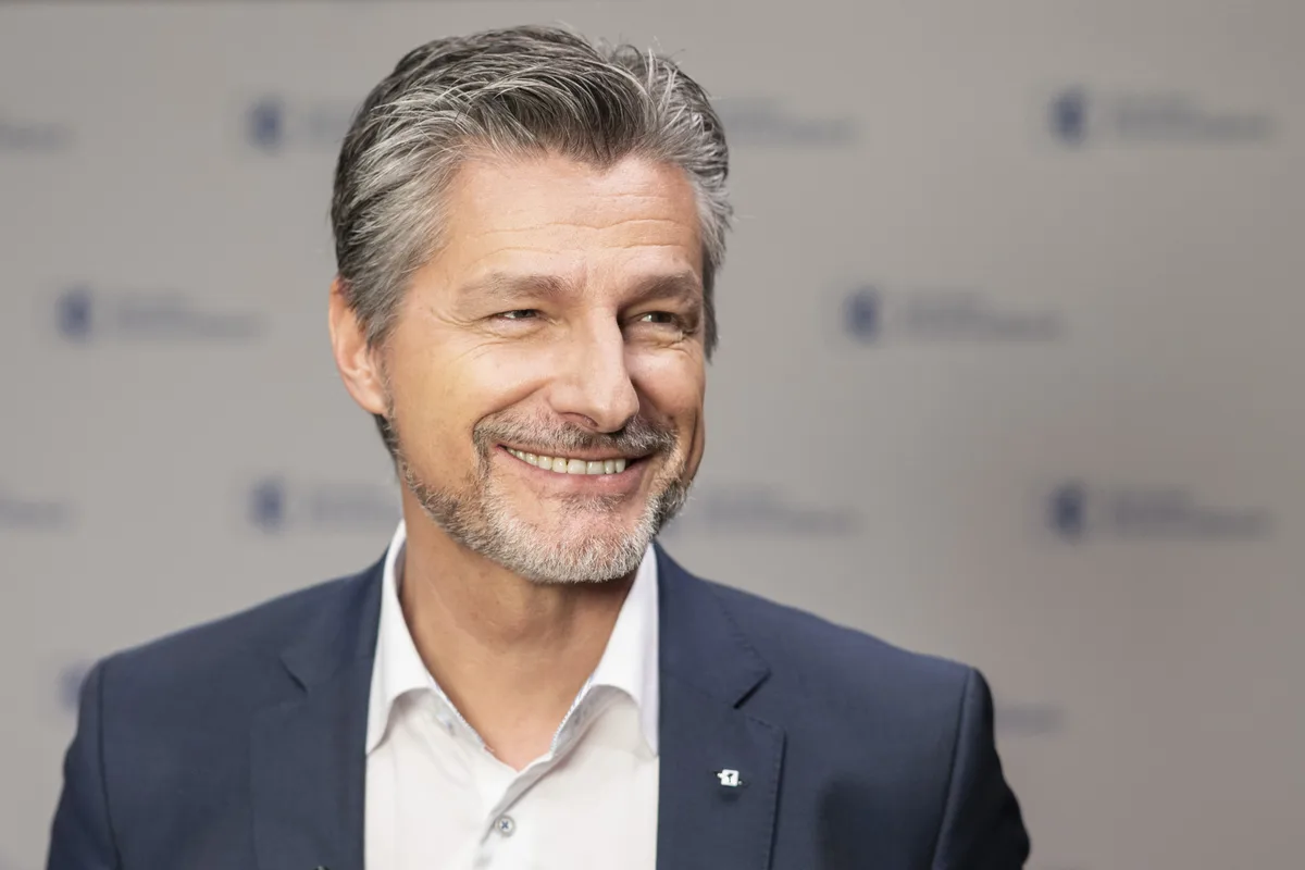 Urs Baumann, CEO der Zürcher Kantonalbank, lächelt in die Kamera. Er trägt einen grau-blauen Anzug und ein Hemd ohne Krawatte.