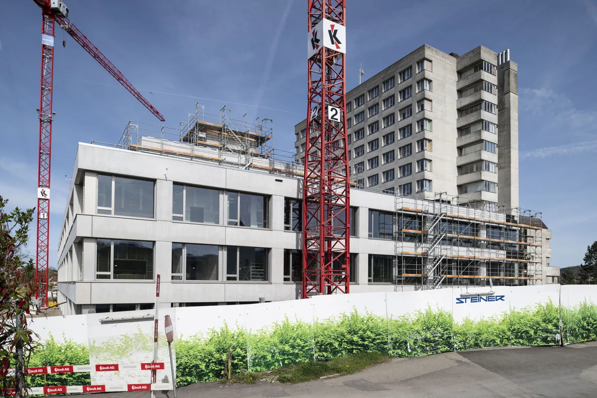 Man sieht ein Gebäude im Bau, davor einen roten Kran, im Hintergrund ein graues Hochhaus.