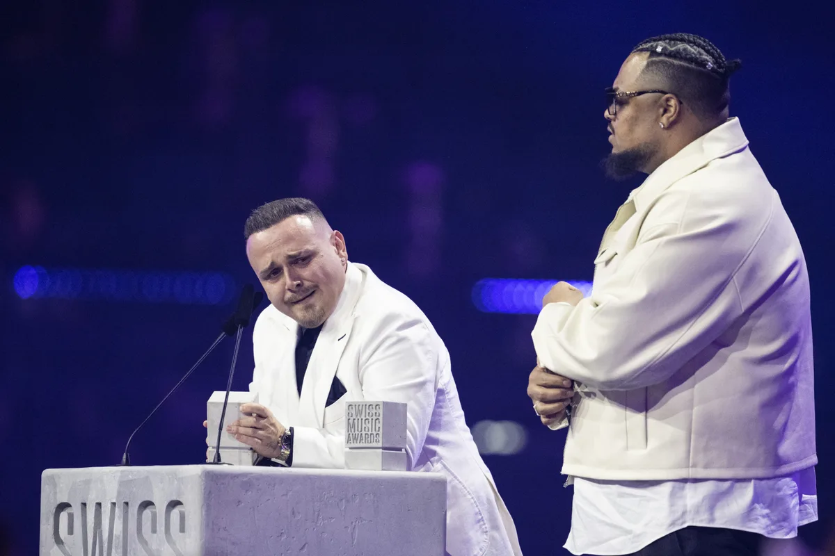 Zwei Männer stehen hinter einem Pult mit Mikrofonen. Beide tragen weisse Jacken.